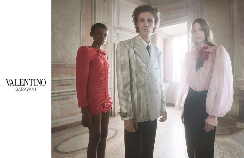 Fashion editorial shot of three models in an elegant, light-filled interior. Two women and one man pose in designer outfits, soft light, and neutral colors.