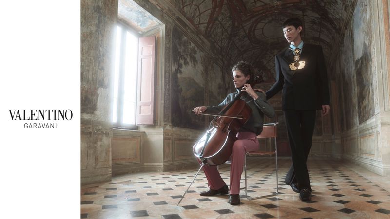 Two fashion models in an ornate indoor setting. One sits playing a cello, while the other stands nearby, wearing a dark suit and pendant.