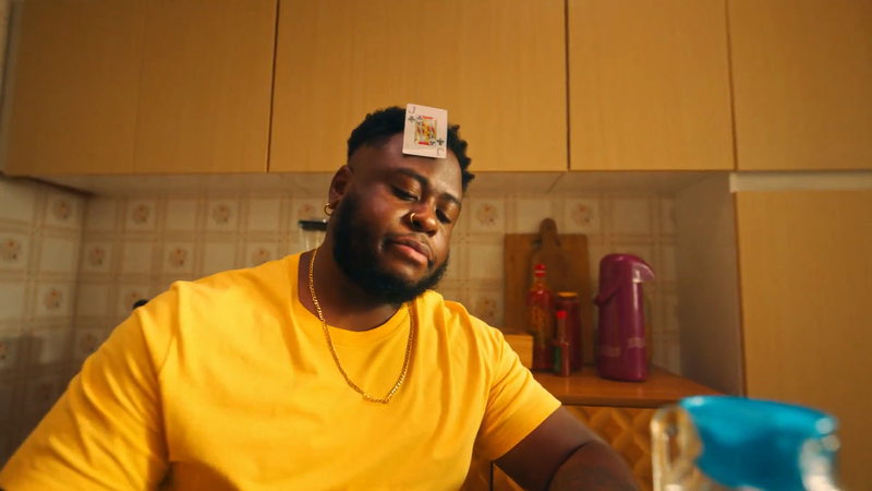 Portrait of a Black man indoors with a Jack of Clubs playing card balanced on his forehead, wearing a gold chain and yellow shirt.