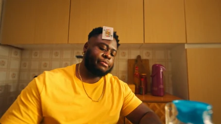 Man wearing a yellow t-shirt and gold chain with a playing card on his forehead in a kitchen