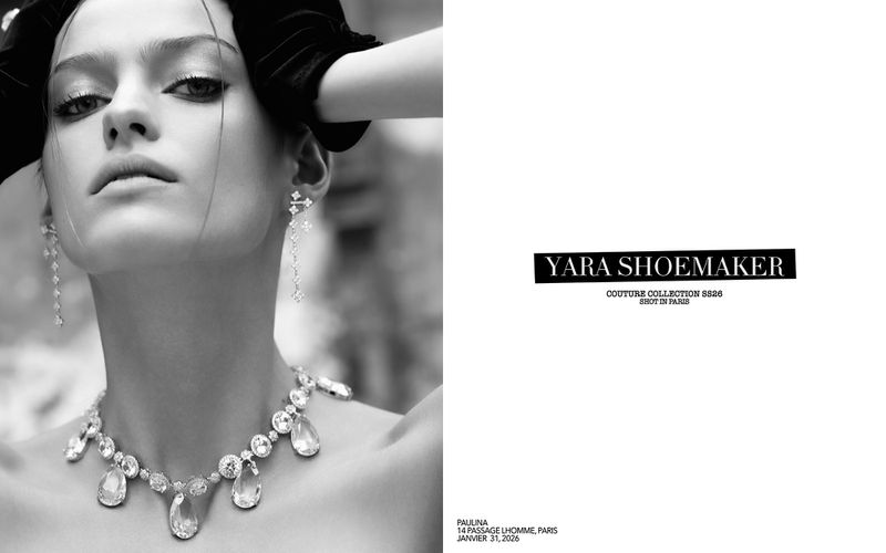 Black and white studio shot of a woman wearing jewelry including a large teardrop necklace, long dangling earrings, and black velvet gloves.