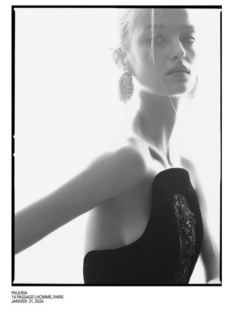 Monochrome portrait of a woman with blond hair wearing a black dress and statement earrings against a white background.