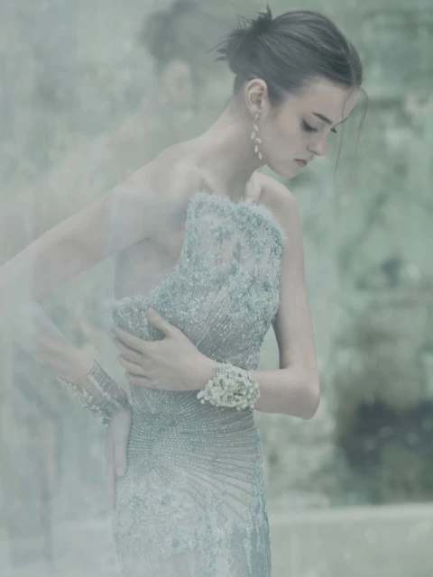 Woman poses in an elegant light blue gown with a one-shoulder design and embellished details, complemented by a cuff bracelet, against a hazy backdrop.