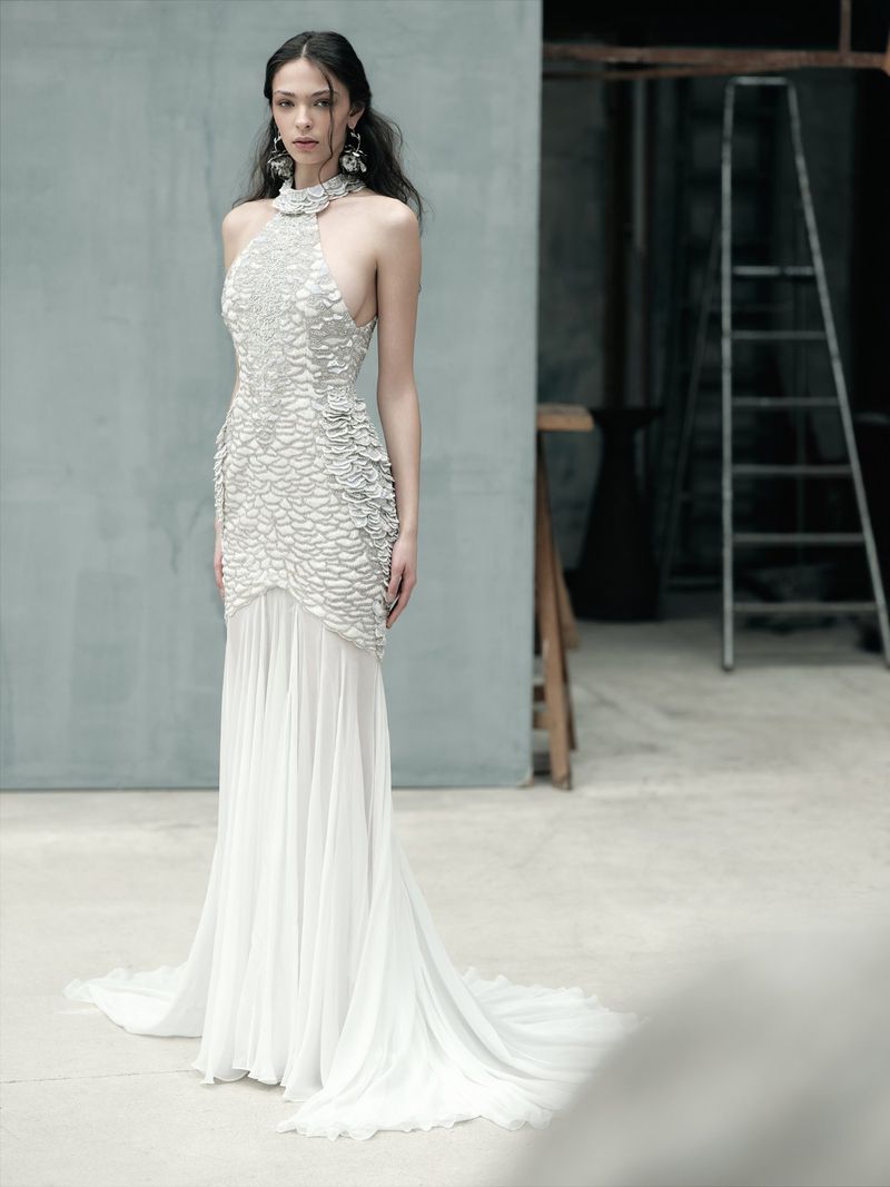 A young woman poses in a couture halter gown with white textured bodice and flowing skirt, standing in a neutral-toned studio environment.