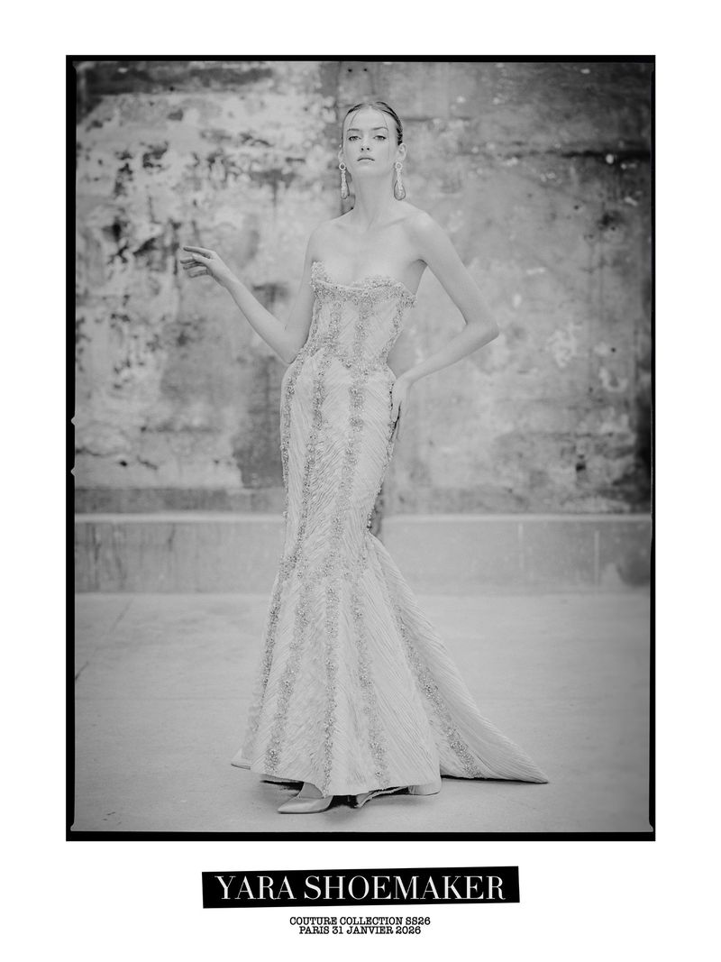 Black-and-white portrait of a woman in an embellished strapless gown with flowing skirt, posing indoors against a mottled wall.