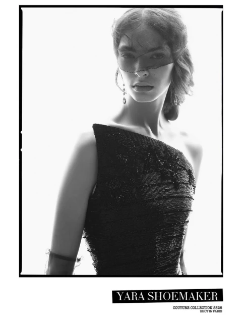 Monochrome portrait of a woman with dark hair and veil, wearing a textured, dark one-shoulder dress against a bright, light background.