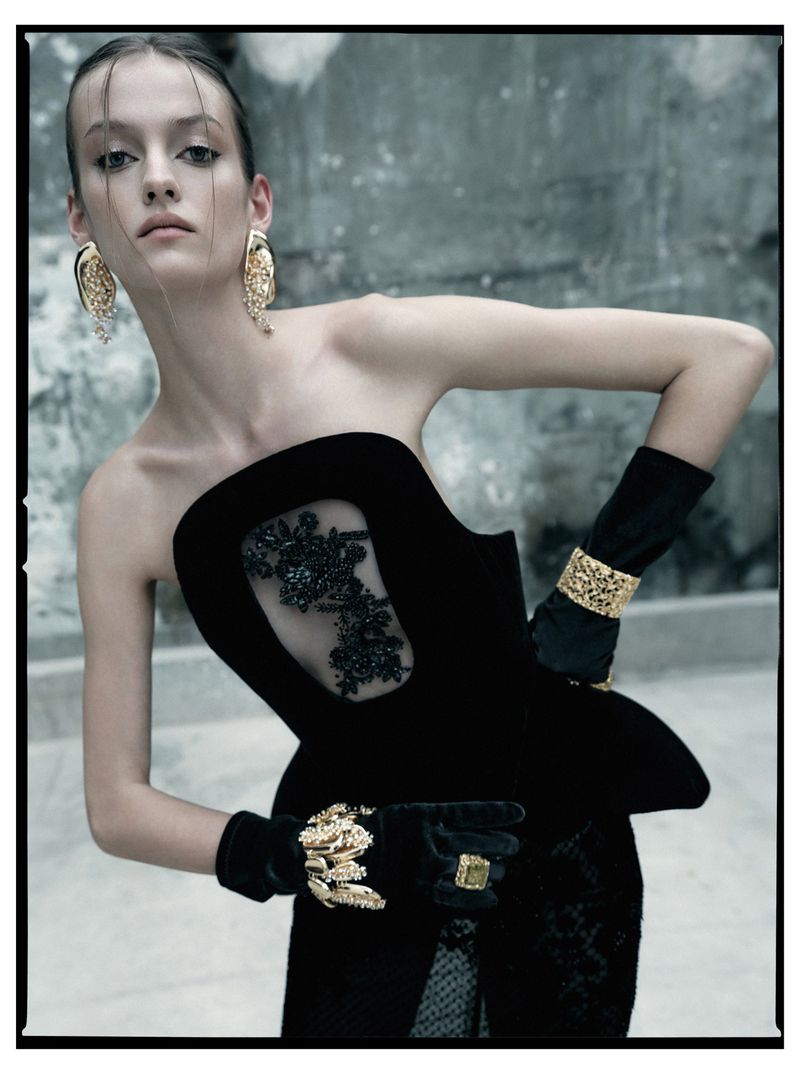Model in black strapless dress with intricate lace cutout, ornate gold earrings and cuffs, standing before a weathered wall.
