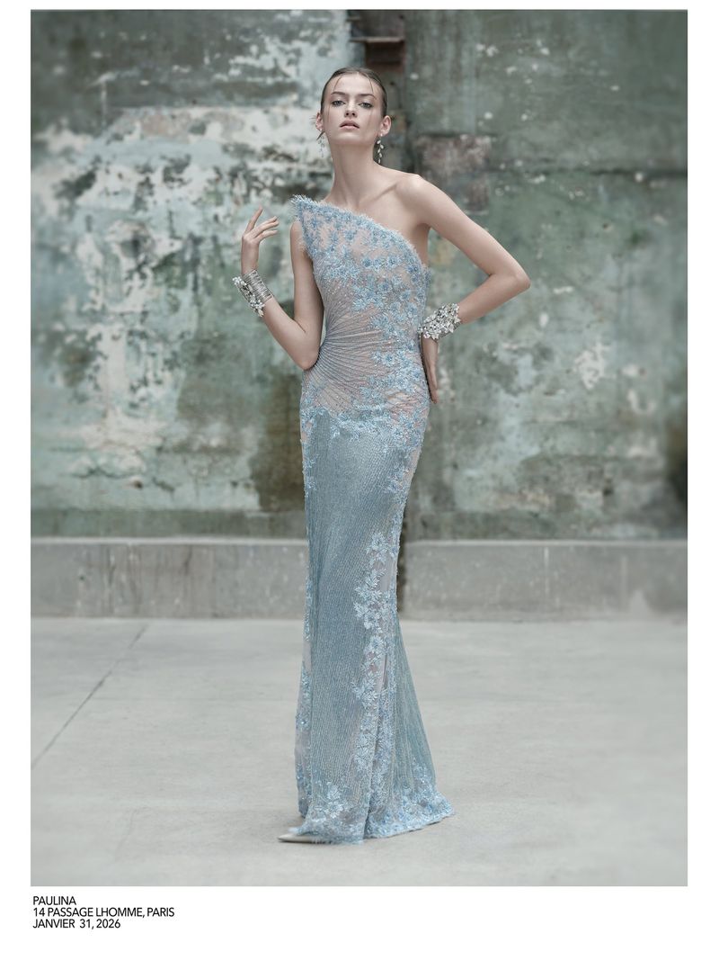 Elegant woman in a light blue, lace-embellished one-shoulder gown posing against a distressed wall in a fashion editorial shot.