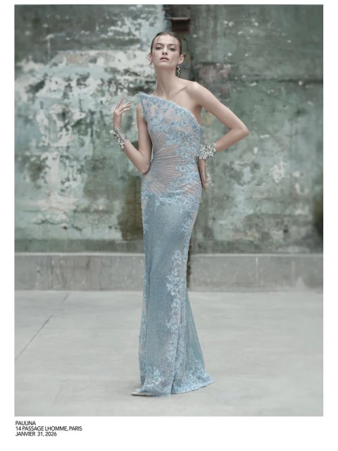 Elegant woman in a light blue, lace-embellished one-shoulder gown posing against a distressed wall in a fashion editorial shot.