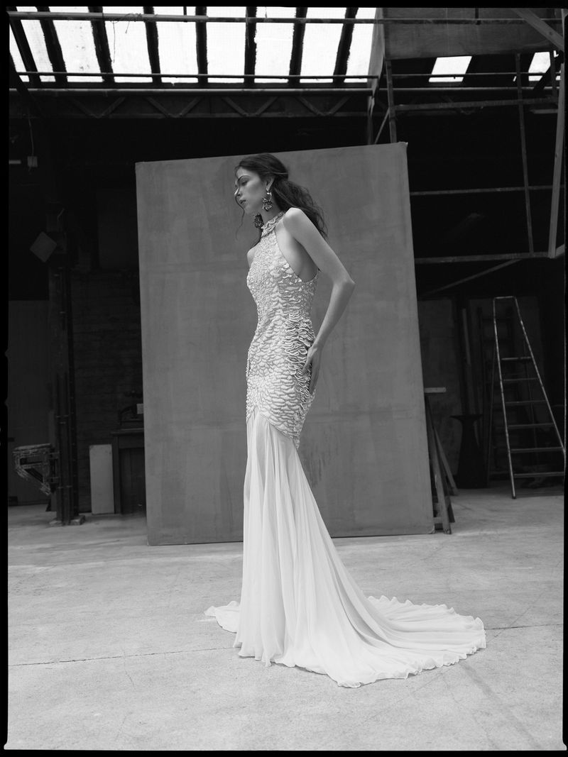 Monochrome portrait of a woman in a beaded gown, standing in a photo studio with soft lighting and visible studio equipment.