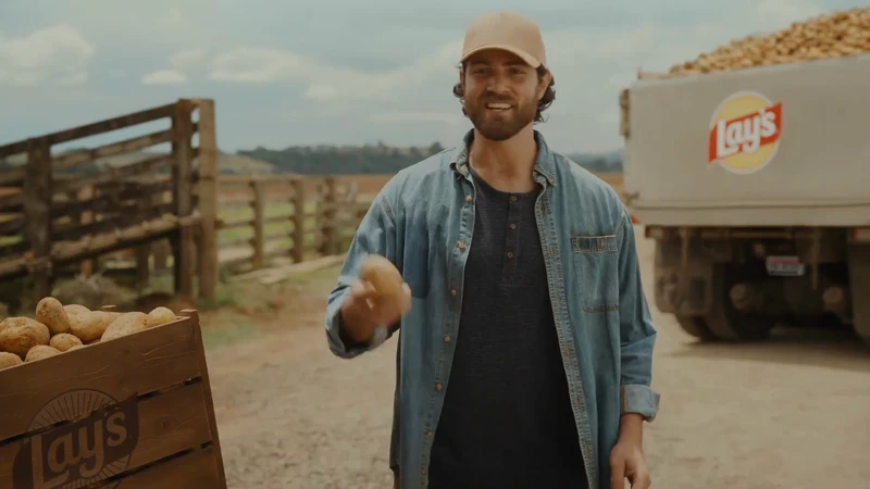 Farmer holds potato in a rural setting with a Lay's potato chip truck visible, showcasing the brand's connection to agriculture.
