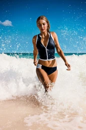 Gigi Hadid wearing a navy and white athletic swimsuit emerging from ocean surf on a sandy beach