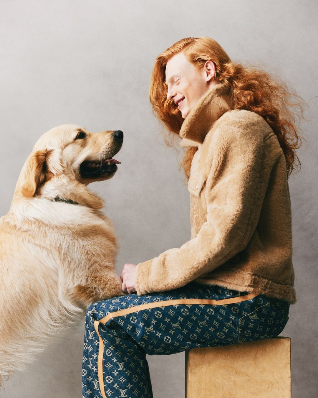 A red-haired adult male smiles and looks at a golden retriever dog in a studio setting, wearing a beige sherpa jacket and patterned pants.