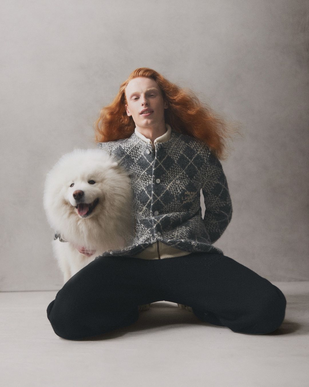 A red-haired male model kneels with a fluffy white Samoyed dog indoors against a grey wall, showcasing a stylish patterned jacket.