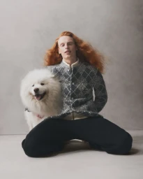 Long-haired red-headed male model kneeling beside a fluffy white Samoyed dog against a neutral grey studio background.