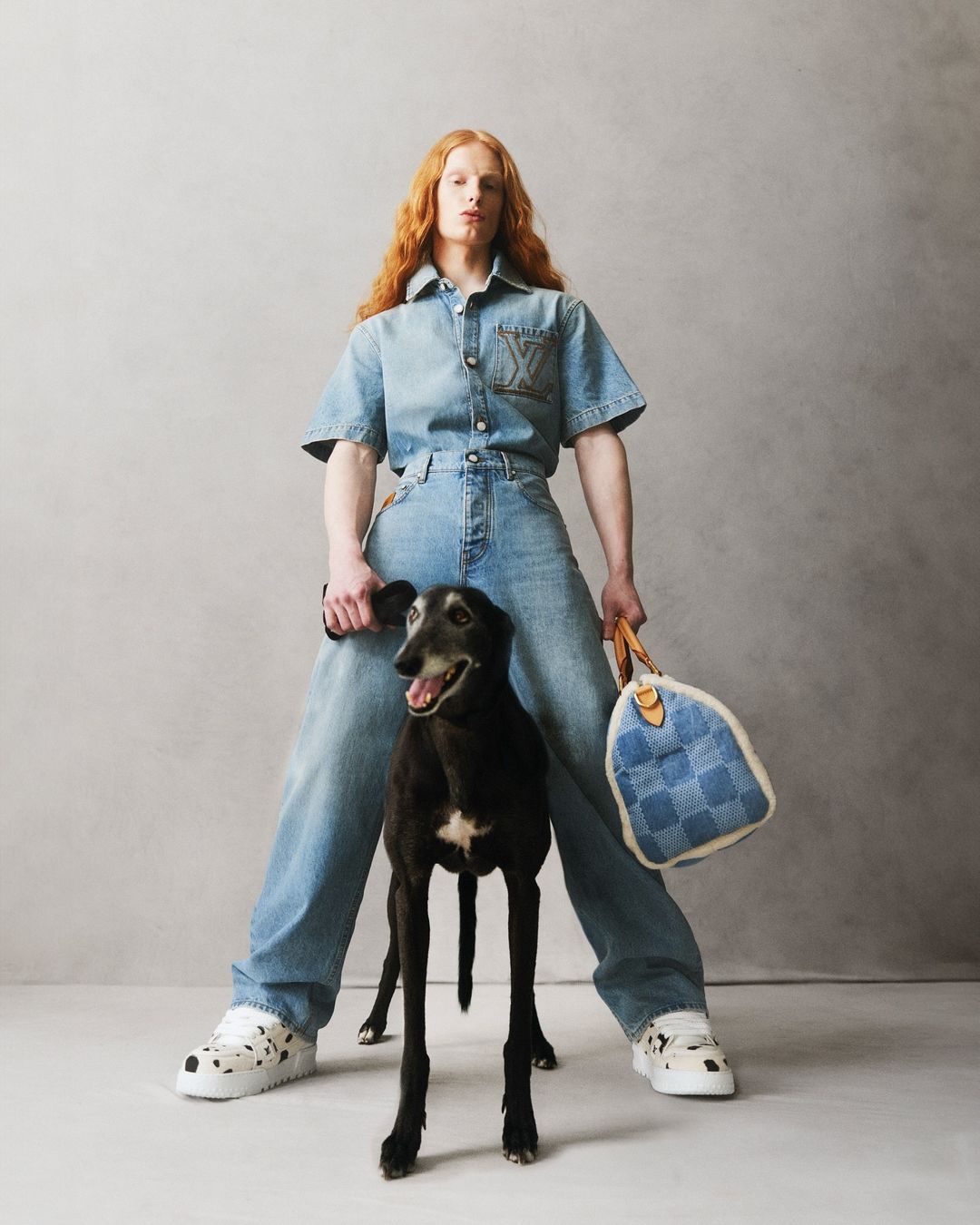 Stylish person in denim outfit poses with greyhound dog and designer handbag against a neutral backdrop in a studio setting.
