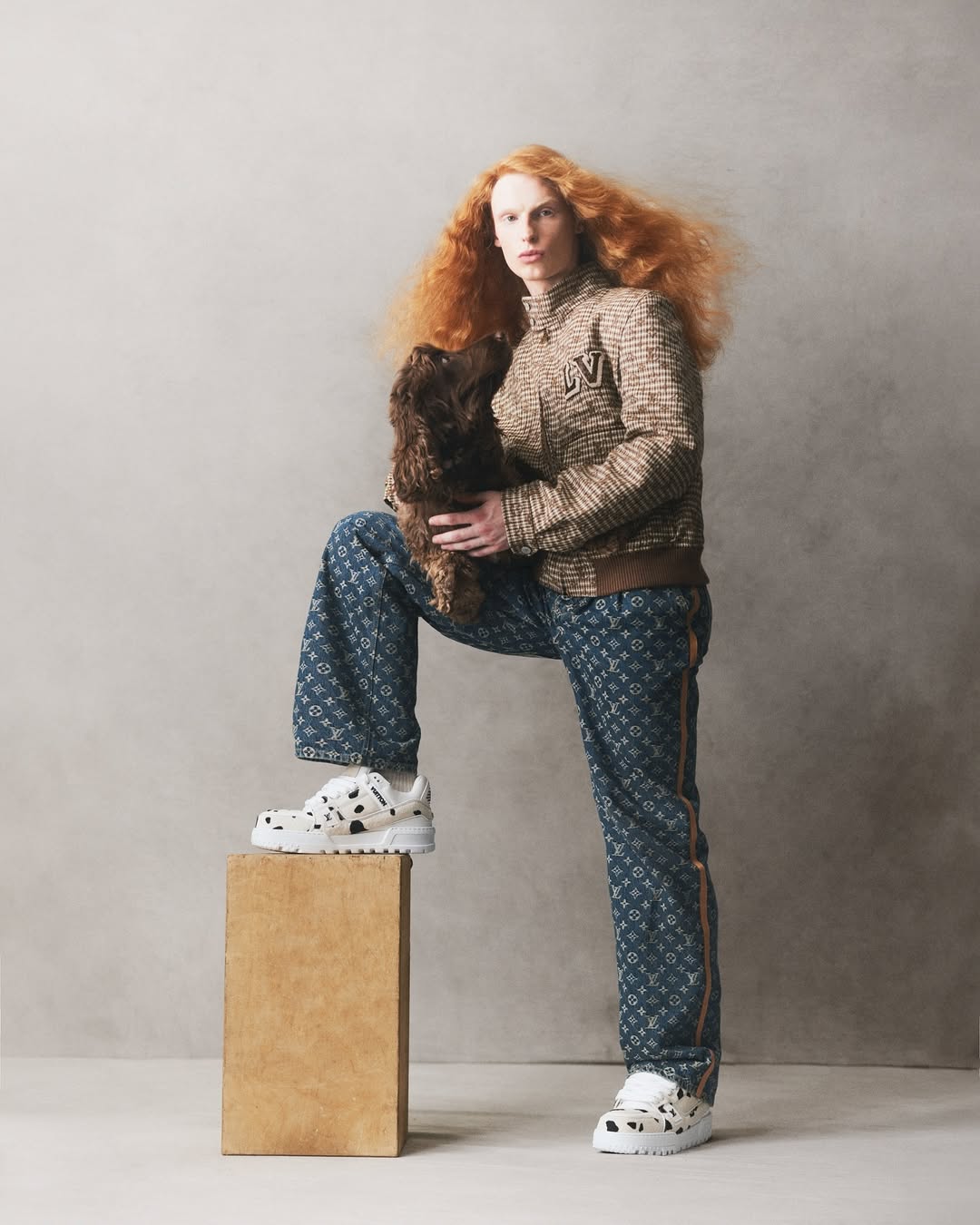 Fashion portrait featuring a red-haired person in patterned outfit with a dog, posing with one foot on a box in a neutral studio setting.