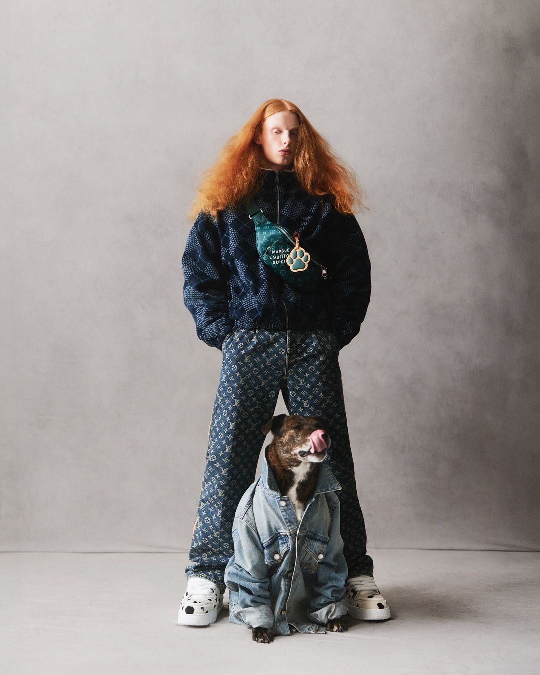 Fashion model with red hair poses in a printed jacket and pants next to a dog wearing a denim jacket indoors on a gray background.