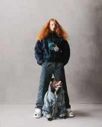 Model with long red hair wearing monogram jacket and pants standing behind dog in a denim jacket against a grey seamless backdrop