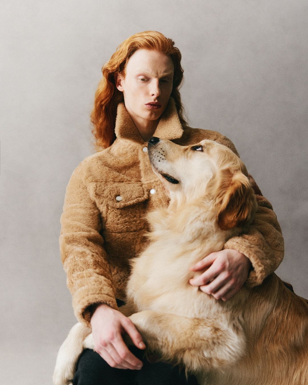 Portrait of a red-haired man in a shearling jacket holding a Golden Retriever dog against a grey backdrop in a photo studio.