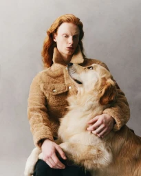 Long-haired man wearing a camel-colored fleece jacket holding a golden retriever dog against a neutral studio backdrop