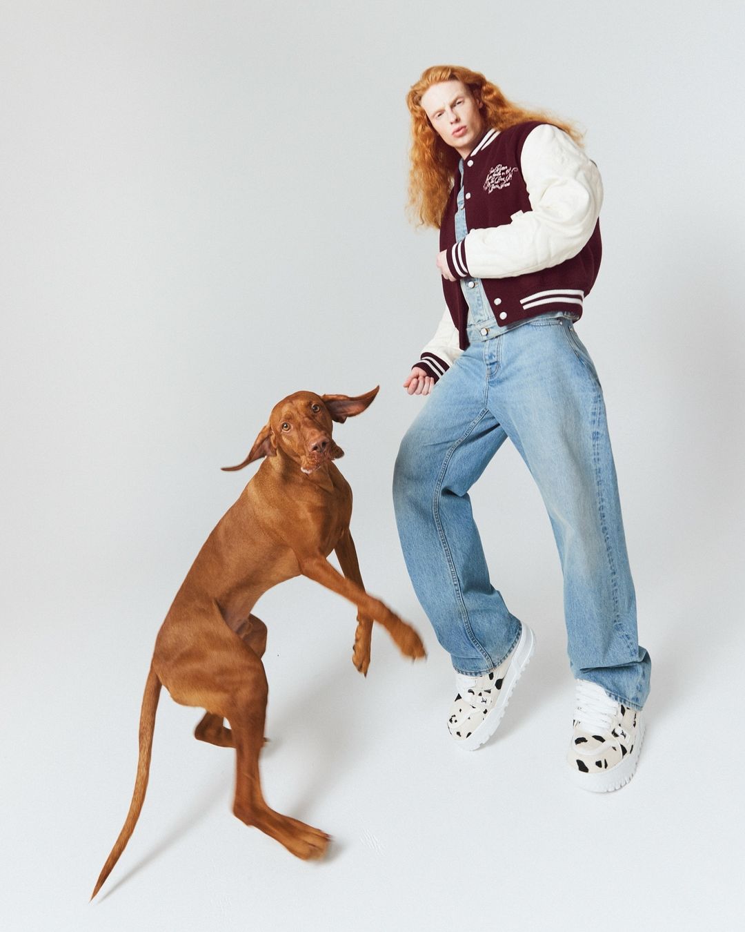 Adult posing with a Vizsla dog in a photo studio. The person is wearing a jacket, denim overalls, and platform sneakers on a white background.