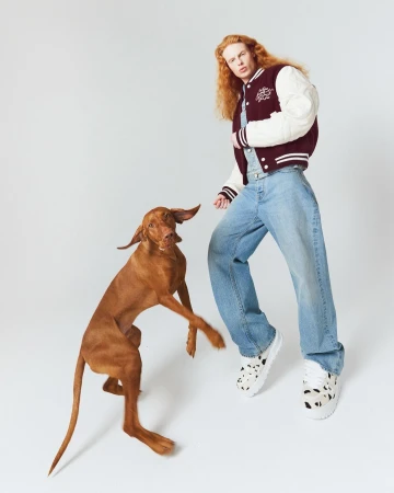 Adult posing with a Vizsla dog in a photo studio. The person is wearing a jacket, denim overalls, and platform sneakers on a white background.