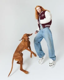 Red-haired man in a maroon varsity jacket and light wash jeans with a brown dog against a white background