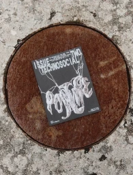 Black booklet with white typography resting on a round, rusty metal manhole cover outdoors.