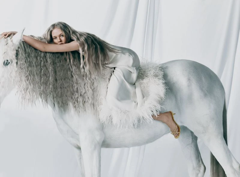 A fair-skinned woman with long platinum hair poses on a white horse wearing matching silk garments in a brightly lit studio.