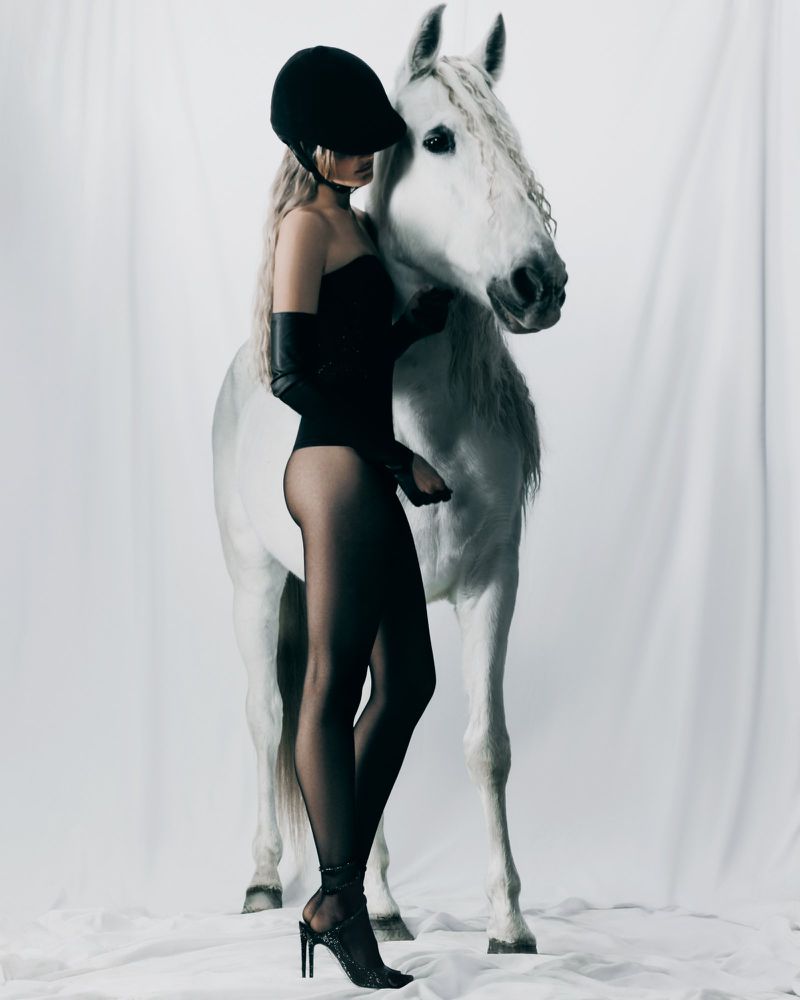 Woman in bodysuit and equestrian helmet poses with a white horse in a studio, capturing a blend of fashion and equestrian aesthetics.