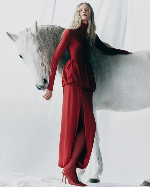Fashion editorial shot of a blond model in a red gown with a horse against a bright white backdrop. High fashion studio portrait.