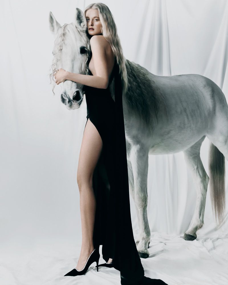 Woman in a black dress posing next to a white horse against a white fabric backdrop in a studio with bright, soft lighting.