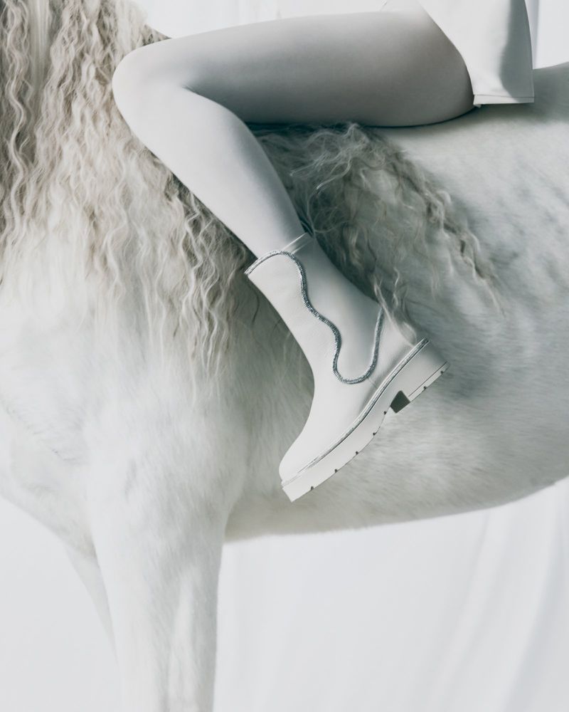 Equestrian fashion concept: a woman in white tights and boots rests her leg on a white horse in a minimalist studio setting.