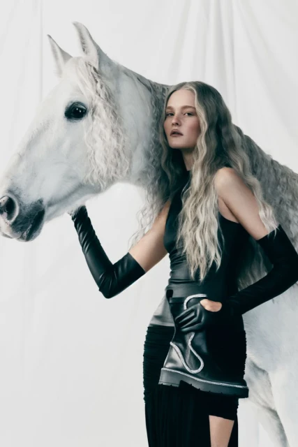 Woman in black dress poses with a white horse in a studio setting, creating a surreal fashion-editorial image with soft lighting.