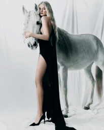 Woman in a black dress standing beside a white horse against a draped white backdrop.