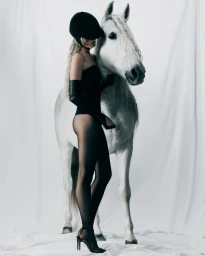 Model wearing a black riding helmet and bodysuit standing beside a white horse against a seamless white background