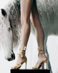 Model standing on a black pedestal wearing gold high heels with floral straps next to a white horse