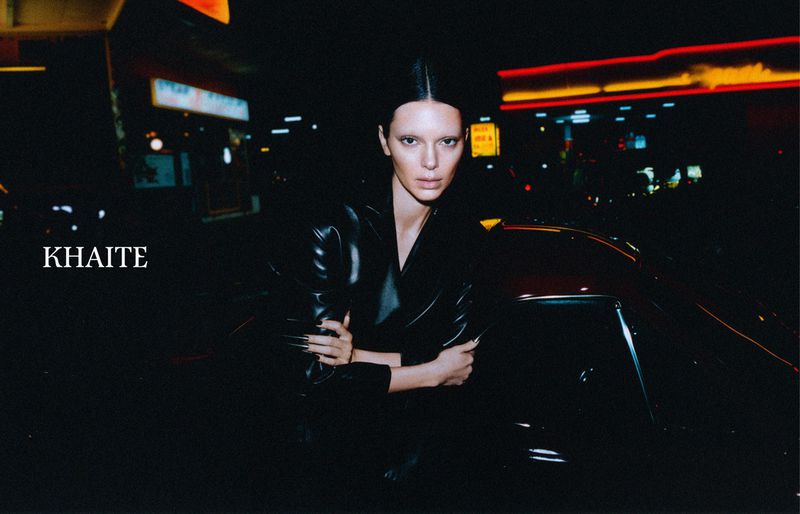 A woman in a black leather jacket poses next to a car at night, with illuminated buildings in the background, in a fashion editorial.