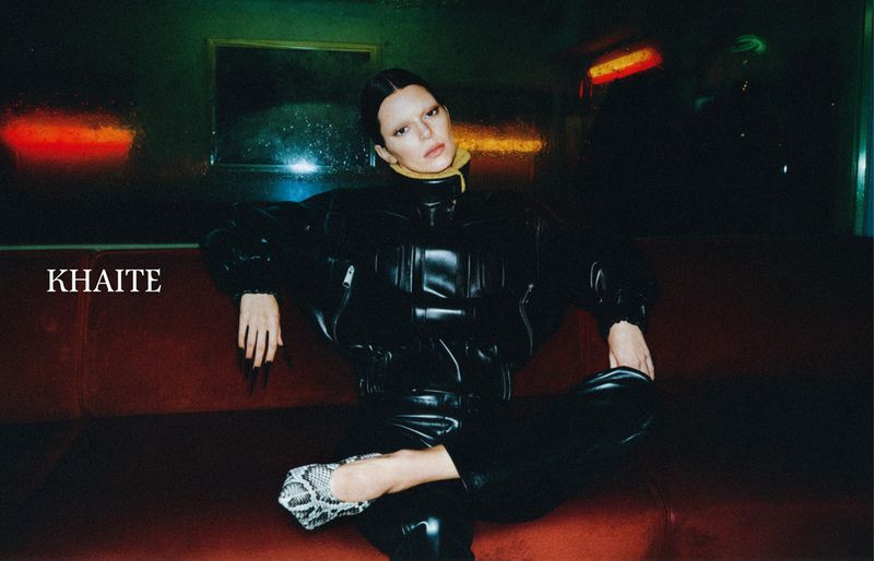 Fashion shot of a woman in black leather clothing reclining on a red sofa in an interior with dark, moody lighting and snake-print shoes.