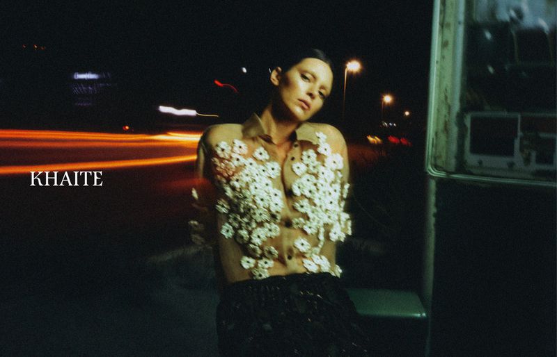 A woman in a sheer floral-embellished top poses at night, the background blurred with car light streaks and an old outdoor telephone booth.