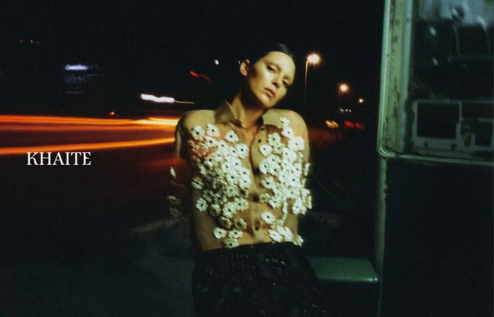 A woman in a sheer floral-embellished top poses at night, the background blurred with car light streaks and an old outdoor telephone booth.