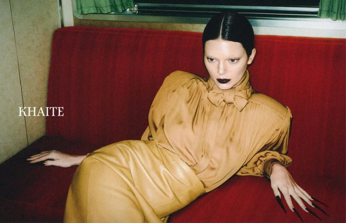 A light-skinned fashion model poses on a red couch in a tan-colored blouse and skirt, with dark lipstick and manicured nails, in a studio setting.