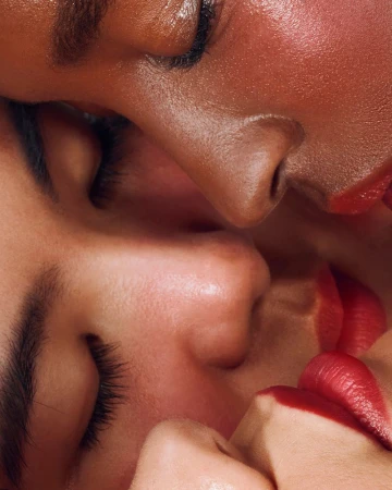 Close-up of two adult females, one Black and one white, with glossy red lips, heads close together and eyes closed, indoor lighting.