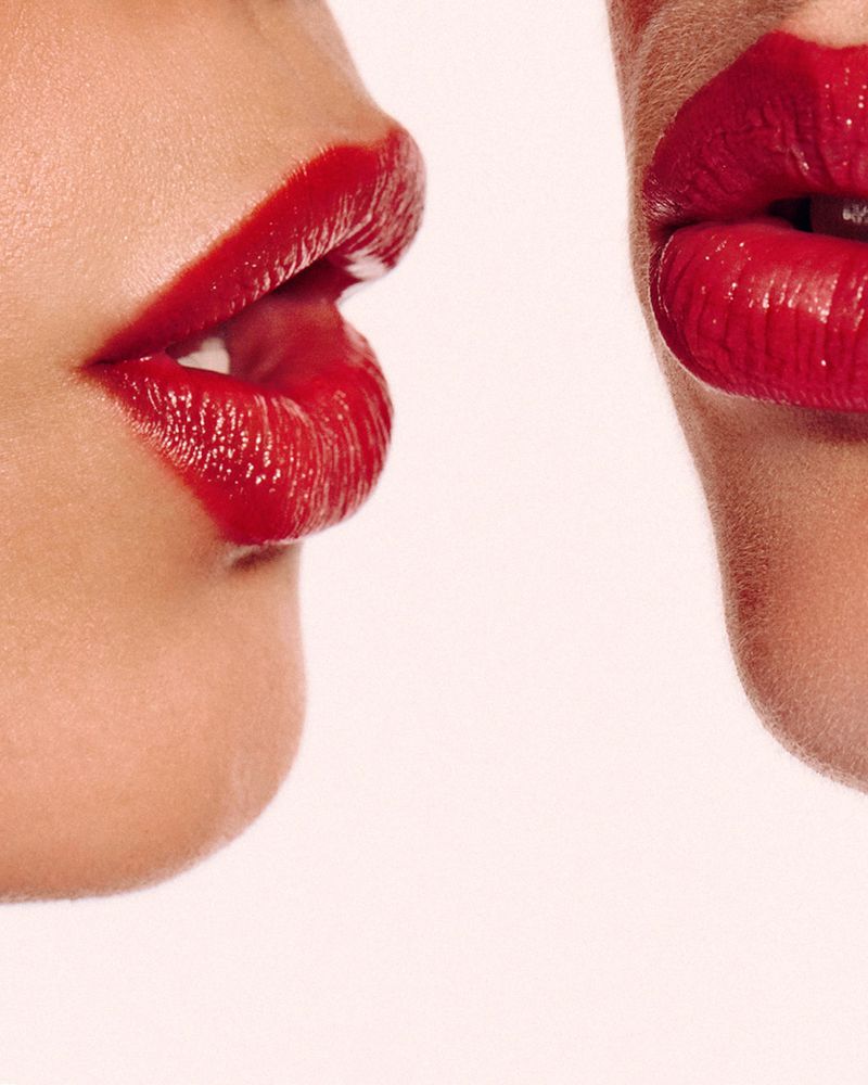 Close-up of two adult females with bright red lipstick. One face is in profile with open mouth, one is facing front with closed mouth.