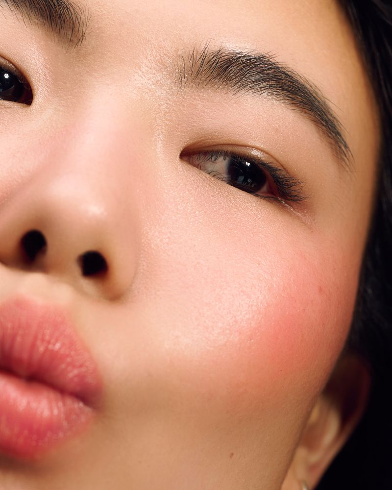 Close-up studio shot of an Asian female's face featuring soft pink lips, dark eyes, fair skin, and pink blush, set against a dark backdrop.