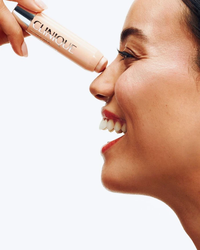 Close-up profile of a smiling woman applying a Clinique Chubby Stick cosmetic product to the tip of her nose.