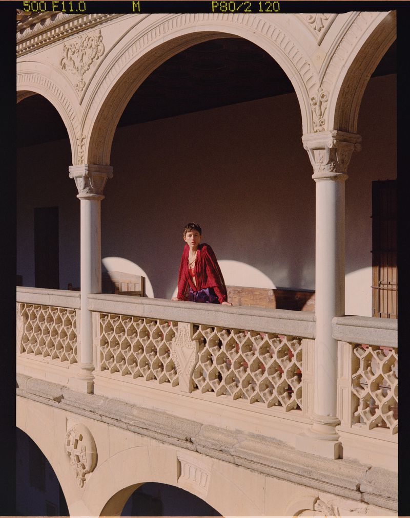 Adult woman in a fashionable red and purple outfit poses on a stone balcony with arches in a sunlit outdoor setting.