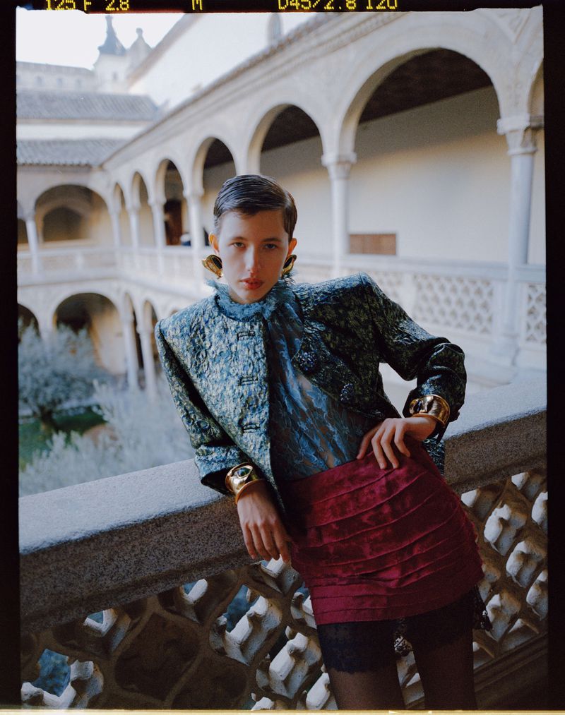 Fashion photo of a person posing in a teal jacket and red skirt on a balcony, overlooking an outdoor courtyard with arched walkways.