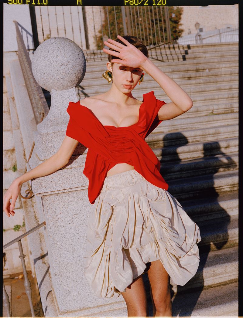 Stylish woman in a red top and cream skirt poses on stone stairs, shielding her eyes from the sunlight in an outdoor fashion shot.
